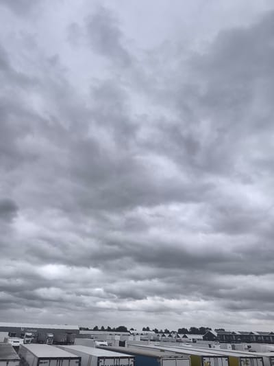 The image depicts an overcast sky filled with dark, dramatic clouds, casting a somber atmosphere over a commercial or industrial area. In the foreground, rows of white semi-trailer trucks, likely parked in a lot or depot, dominate the lower half of the frame. Some of the trailers show branding or logos, although they are difficult to read clearly due to the distance and lighting. Behind the trucks, a long, low building with the word "FLOWERS" visible above an entrance is partially obscured. Further in the background, a line of smaller buildings with pitched roofs, resembling housing or smaller commercial units, stretches across the landscape, bordered by trees. The overall scene suggests a logistics or distribution hub, possibly on the outskirts of Uithoorn, Netherlands, given the characteristic architecture of the smaller buildings. No people are visible in the image.