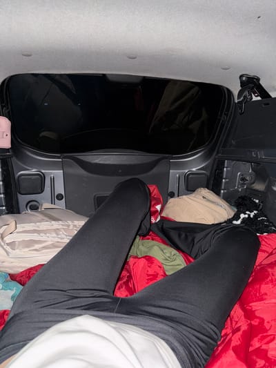 The image shows a first-person perspective inside the cargo area of a vehicle, likely a car or SUV, with the tailgate open and facing outwards into darkness. The interior of the cargo area is filled with various items, including bedding (red and green fabrics), bags (light beige and black), and what appears to be a dark, fuzzy blanket or rug. A person, only their legs and lower torso are visible, is lying down amongst these items, wearing black leggings and a white shirt. Their legs are extended, with the left leg bent at the knee and the right leg mostly straight. The scene suggests a temporary sleeping or resting arrangement within the vehicle. There is no visible context suggesting a specific location within the city of Lasva, Russia, or any urban environment; the background is entirely dark.