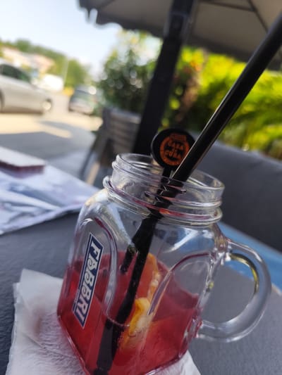The image shows a close-up of a red drink in a mason jar glass. The glass has a handle and a logo that reads "FABBRI 1905". A black straw is inserted into the drink, and a small circular tag with "CASA de CUBA" written on it is attached to the straw. A slice of orange or lemon is visible within the drink. The glass is resting on a dark surface, possibly a table, with a white napkin underneath. In the background, out of focus, are trees, a white car, and what appears to be a building with a light-colored facade. The scene suggests an outdoor seating area, potentially a cafe or restaurant, on a sunny day. The presence of the "CASA de CUBA" tag might indicate a themed drink or establishment. Without further information, it is impossible to determine the specific location within Plovdiv, Bulgaria.