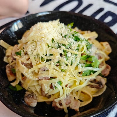 A close-up shot showcases a black bowl filled with fettuccine pasta, generously topped with shredded cheese and chopped parsley. The pasta appears to be mixed with pieces of bacon and mushrooms, coated in a creamy sauce. The bowl rests on a patterned tablecloth. The image is focused on the food, with a blurred background that includes a hint of a person's shoulder in the upper left corner. The scene is indoors, likely a dining setting in Locking Parklands, United Kingdom.