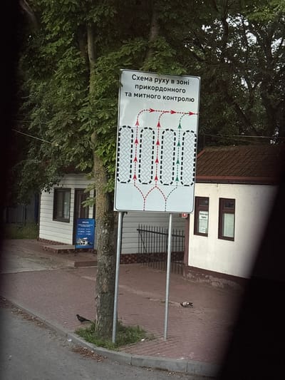 The image shows a street scene in Mohyliv-Podilskyi, Ukraine, with a traffic sign and a building. The traffic sign, in Ukrainian, displays a diagram of traffic flow in a border control zone. It has red arrows indicating traffic lanes and green arrows indicating the direction of entry. The sign is located next to a tree on a paved walkway. To the right of the sign is a white building with brown-trimmed windows. One window has a poster with text in Cyrillic, possibly advertising services. The ground is paved with red bricks. A pigeon is visible on the walkway. The overall environment suggests a public area, possibly near a border crossing or administrative building.