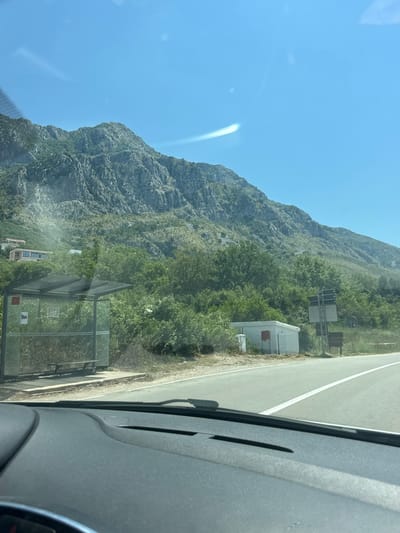 The image captures a view from a vehicle dashboard, looking out towards a rural road scene in Blizikuće, Montenegro. The foreground shows the dark grey dashboard and windshield. In the mid-ground, a paved road runs horizontally across the frame, with a white lane marker. To the left of the road is a bus stop shelter with a transparent roof and glass sides, containing a wooden bench. A red sign is visible on the side of the shelter. Adjacent to the bus stop is a small, white, box-like structure with a red mailbox on its side. Further to the right, a metal pole with signage stands near the road. Behind these elements, a dense green forest covers a sloping hillside. Dominating the background is a large, rugged, grey mountain range, with its rocky slopes partially covered by vegetation. The sky is a clear, bright blue with a faint, horizontal streak of light across it. A few modern buildings with large windows are visible on the upper part of the hillside, nestled among the trees. The overall impression is a sunny day in a mountainous, coastal region.