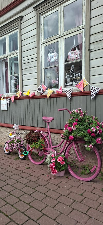 The image depicts a charming storefront in Viljandi, Estonia, with a focus on the exterior decorations. Two vintage-style bicycles, painted pink, are prominently displayed. The larger bicycle is laden with a lush arrangement of pink and white flowers in its basket and also has plants in a pot on its rear rack, with more flowers placed at its base. A smaller pink bicycle is positioned to the left, also adorned with pink flowers and a whimsical yellow duck toy on its handlebars. The bicycles are placed on a cobblestone pathway.
Behind the bicycles, the building features weathered wooden siding painted a muted grey, with classic wooden window frames. The windows are large, with multiple panes, and reflect the overcast sky. Inside the windows, two floral-patterned handbags are visible, suggesting the nature of the shop, possibly selling handmade goods or accessories. A string of colorful triangular pennants, featuring polka dots and floral patterns in pink, yellow, and black, is draped across the front of the building above the bicycles. Signage visible on one of the windows, though partially obscured, includes text that appears to be "De la Rose" and "KÄSITÖÖKOTID", indicating the shop's name and possibly that it sells handmade bags. Another sign on the left window reads "LAATUD OPEN". The overall scene conveys a sense of quaint, decorative storefront display, likely intended to attract customers and add to the aesthetic appeal of the street in Viljandi. There are no people visible in the image.