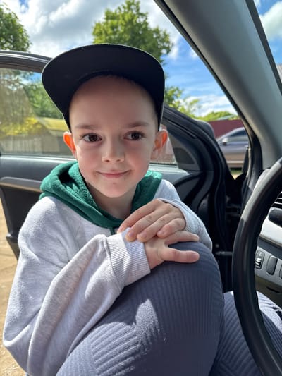 A young boy wearing a dark baseball cap and a grey hooded sweatshirt with green trim sits in a car. The open car door is visible. A steering wheel is partially visible in the background. Outside the car, trees and a blue sky with clouds are seen. The image does not provide information about the location of Slobodka, Russia.