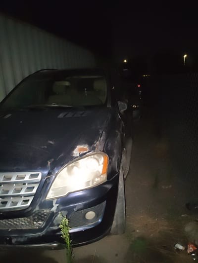 A dark-colored SUV is parked at night next to a corrugated metal wall on the left and a chain-link fence on the right. The SUV's headlights are on, illuminating the front of the vehicle and the ground. The vehicle appears to have some damage, with rust visible on the hood and bumper. The area around the vehicle is unkempt, with dry grass and debris on the ground. In the background, there are streetlights visible, suggesting the location is within a city. Based on the provided information, this scene is located in Krtsanisi, Georgia.