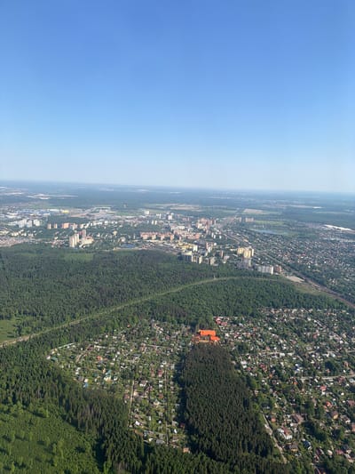 The image shows an aerial view of Перепечино, Russia, with a clear blue sky above. The landscape transitions from a dense forest in the foreground to a mix of residential and urban areas in the distance. Patches of greenery are interspersed throughout the city, and a railway line runs parallel to a large forest. An orange building is visible amidst a cluster of houses in the residential area. The environment appears to be affected by some amount of haze or smog in the distance.