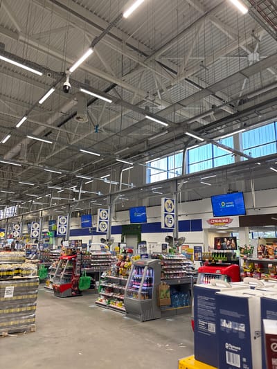 This media file captures the interior of a large hypermarket, likely in Dimitrovgrad, Russia, oriented to show the extensive ceiling structure and multiple shopping aisles. The scene is brightly lit by numerous fluorescent tube lights and natural light filtering through skylights in the high, industrial-style ceiling, which features exposed metal beams, trusses, and ventilation ducts. Surveillance cameras are also visible on the ceiling infrastructure.
The core activity is retail shopping, evidenced by the clearly marked aisles and stocked shelves. Blue and white signs hang from the ceiling, indicating aisle numbers from 6 to 15, guiding customers. Digital displays overhead prominently feature the "гипер лента" (Hyper Lenta) logo. Another significant sign advertises "СТАНЦИЯ СТРИТФУД" (Station Street Food) and "СЕТЬ КАФЕ БЫСТРОГО ПИТАНИЯ" (Network of fast food cafes), suggesting a food service area within the store.
Along the visible aisles, refrigerated display cases contain beverages, and numerous pallets of bottled water are stacked on the floor. Shelves are stocked with various products, including snacks like chips, and other packaged goods. A red shopping cart is partially visible near a display. Text such as "Оставьте отзыв:" (Leave feedback:), "100-", "3x", and "ETIB PARFUM" can be seen on various labels and signs. The floor is a light, polished concrete. The setting is clean, organized, and appears to be bustling with merchandise, though no specific individuals are clearly visible in the frame. The overall impression is that of a modern, well-maintained supermarket environment during daytime hours.