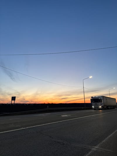 The image depicts a roadside scene at dusk or dawn, possibly in the vicinity of Kuregovo, Russia, given the presence of a truck with the text “ГЗК Пермскии край” (GZK Perm Region) visible on its side.
A large, white semi-truck is positioned on a multi-lane road. The road surface is asphalt and features lane markings. Streetlights illuminate the scene, one of which is positioned just before the truck.
The sky transitions from blue to orange/yellow, suggesting a sunset or sunrise. A strip of trees or vegetation is visible along the horizon. Power lines stretch across the sky in the left portion of the image.