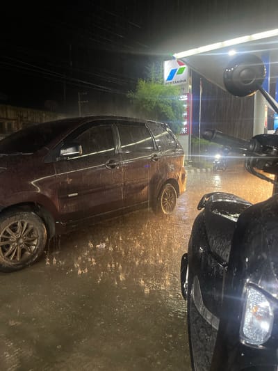 At night, during a heavy downpour, two vehicles are visible at a Pertamina gas station in Barat Yaman, Indonesia. A dark red van is parked on the left side of the frame, its surface slick with rain. In the foreground, the right side of a black motorcycle is visible, also wet from the rain. The ground is flooded and reflects the light from the gas station canopy and streetlights, creating an orange glow. Raindrops are clearly visible in the air and on the surfaces of the vehicles. The Pertamina logo is prominently displayed on a sign behind the van. The scene depicts a common occurrence during the rainy season in Indonesia.