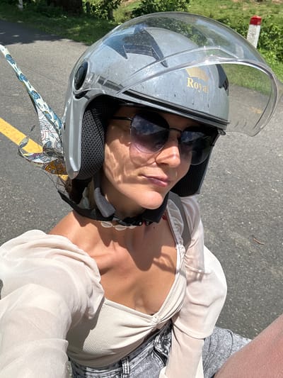 A woman wearing a grey helmet with a clear visor and sunglasses rides a scooter on a paved road. A patterned scarf is attached to the helmet. She is wearing a cream-colored top with puffed sleeves and grey shorts. The location is Ham Thuan Bac, Vietnam, a rural area with trees and vegetation visible in the background. The road has a yellow line marking the lane.