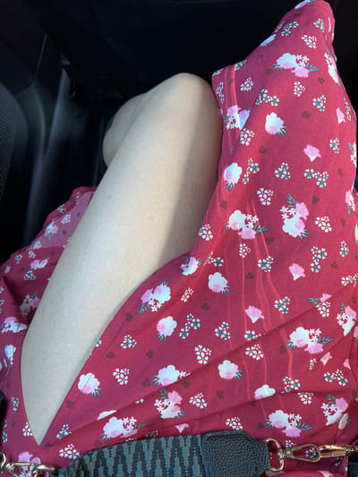 The image depicts a person's bare leg and a portion of a red floral dress, seen from an elevated perspective within what appears to be a vehicle's interior, in Cassano delle Murge, Italy. The central focus is on a fair-skinned leg, likely the thigh and knee area, positioned on the left side of the frame. The skin exhibits a smooth texture with minor visible hair follicles and subtle, lighter discolorations. Covering the lower and right portions of the frame is a red dress featuring a repeating pattern of stylized white and pink flowers, some with green accents, along with smaller dark brown shapes. Distinct parallel rays of sunlight are visible across the fabric of the dress. In the bottom right corner, a black and grey patterned strap, secured by a metallic gold-colored clasp, is partially visible, suggesting a bag. The background is dark and indistinct, consistent with the shadowed interior of a car, with a textured black surface on the far left, possibly an upholstered seat or door panel.