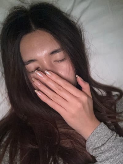A close-up image depicts an individual, likely female, lying down with eyes closed. Her dark brown hair, parted centrally, spreads around her head. Her right hand, adorned with long, almond-shaped fingernails painted with a pearlescent polish showing visible regrowth, covers her nose and mouth. She wears a grey long-sleeved garment on her right arm. The background consists of a plain, light-colored, soft surface, consistent with bedding or a pillow, indicating an indoor setting.