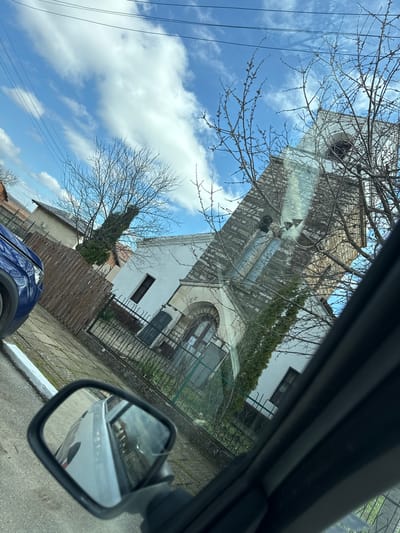 The image, taken from inside a car, shows a view of an old church in Kameno Pole, Bulgaria. The church is made of stone and has a tall bell tower. A tree with bare branches stands in front of the church, partially obscuring the view. The sky is blue with white clouds. The car's side mirror reflects a street scene with buildings. A portion of a blue car is visible at the bottom of the image. The image is tilted at an angle. There are utility lines visible in the sky. A wooden fence surrounds part of the property.