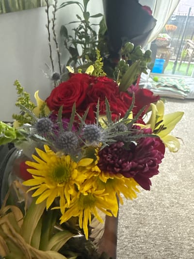 A floral arrangement, featuring red roses, yellow sunflowers, purple carnations, and yellow lilies, is displayed indoors. The backdrop includes a window reflecting an outside scene and a textured wall. The arrangement is in a glass container. There is no indication of the location being Des Plaines, United States.