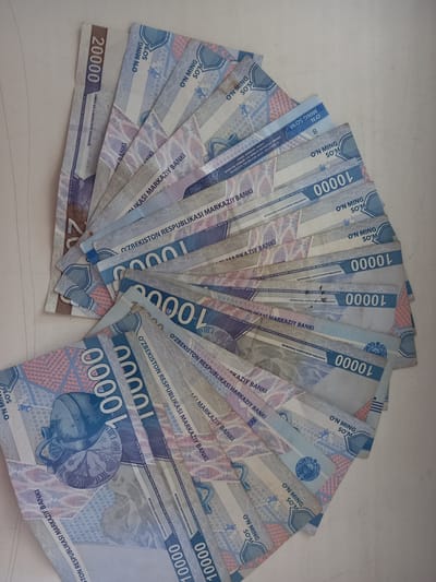 The image shows a fanned arrangement of Uzbekistani Som banknotes, predominantly 10,000 Som and some 20,000 Som denominations. The banknotes are predominantly light blue and white with intricate patterns and emblems. The image is a close-up, top-down view of the currency spread out on a light-colored, plain surface, likely a table or countertop. There are no people visible in the image. The lighting appears to be ambient, casting soft shadows. The overall impression is of a neat presentation of currency. The image was taken in the city of Keles, Uzbekistan.
