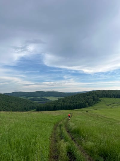 An adult, wearing an orange shirt and dark shorts, runs away from the camera on a dirt path through a grassy field. The path winds through rolling green hills covered in dense forest. The sky is cloudy with patches of blue visible. The image appears to be taken in Zykovo, Russia.