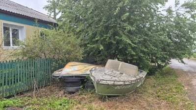 In Timluy, Russia, two old, weathered boats are parked side-by-side in a grassy yard next to a wooden fence. The boat in the foreground is green, with a partially transparent windshield. The other boat, yellow and blue, is propped up on tires. Behind the boats, a large, leafy tree casts a shadow, and a portion of a yellow and teal house with a gray roof is visible. The ground is covered in dry grass and weeds, and a dirt road is visible to the right of the scene. The sky is overcast.