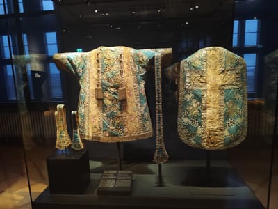 The scene captures three richly embroidered ecclesiastical vestments displayed within a clear glass case inside a museum in Aschaffenburg, Germany, likely the Stiftsmuseum der Stiftskirche St. Peter und Alexander.
The central subjects are a dalmatic or tunic and a chasuble, accompanied by two smaller fabric liturgical pieces. The garment on the left is a dalmatic, a short-sleeved vestment with open sides, displayed on a dark, slender stand. It is made of intricate fabric featuring vertical gold-embroidered bands adorned with tassels and floral motifs, interspersed with teal-blue sections patterned with a leopard-spot design and additional floral embroidery. A gold fringe borders its lower edge. To its right, the chasuble, a rounded, poncho-like outer vestment, is displayed from the rear, also on a dark stand. Its back features a prominent gold-embroidered cross design, richly decorated with floral and geometric patterns, set against the same teal-blue and leopard-spot fabric. To the left of the dalmatic, two smaller, similarly ornate fabric strips, possibly manipules or stoles, are presented on miniature dark stands.
All items are set against a dark background within the display case. A dark rectangular information plaque, with white text partially visible (referencing "Orden der Monsignore..." and "1779"), rests on the dark display base in front of the dalmatic. Reflections on the glass display case reveal a large, multi-paned window with internal blinds or coverings, a green emergency exit sign, and general ambient light from the museum environment. No individuals are depicted as subjects in the scene, nor are any actions or direct interactions observable.
