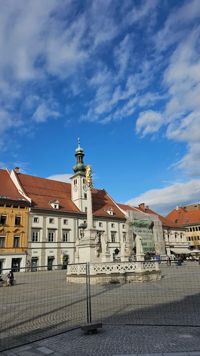 The image depicts the Main Square (Glavni trg) in Maribor, Slovenia, featuring the Plague Column, also known as the Monument to the Holy Virgin. The column is a prominent white structure with a golden statue on top, likely representing the Virgin Mary. Buildings with orange tile roofs and light-colored facades frame the square. A paved pedestrian area stretches out in front of the monument. The sky above is a bright blue with scattered white clouds. People are visible walking in the background.
