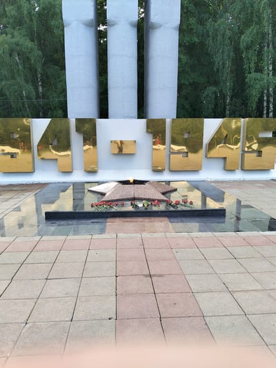 The image shows a war memorial with the inscription "1941-1945" in large, golden, three-dimensional numerals against a white wall. In front of the numerals, a perpetual flame burns over a star-shaped monument adorned with red carnations. The monument is situated on a reflective surface, possibly polished black granite, which is surrounded by paving stones. Behind the memorial, three large, white, pillar-like structures stand tall. The background consists of dense green trees, suggesting an outdoor setting in a park or public space. The overall scene is solemn and commemorative, likely located in Nevzorovo, Russia, as indicated by the prompt. There are no people visible in the image.