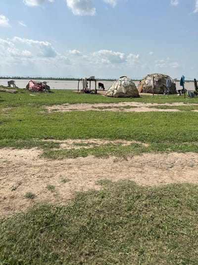 This image depicts an outdoor scene in Dobeli, Nigeria. The subjects are primarily two large, dome-shaped structures that appear to be makeshift dwellings, constructed from dried grasses and possibly fabric. Around these structures, several people are visible, along with various objects that suggest daily life, such as clothing, bags, and possibly a metal table and chairs. A black animal, likely a goat or small cow, stands near one of the dwellings. In the background, a body of water stretches towards the horizon, with a distant line of trees or vegetation. The ground is a mix of dry, sandy earth and green grass. The sky is a light blue with scattered white clouds. The overall impression is one of a rural, possibly temporary, settlement. The lighting suggests it is daytime, likely late morning or early afternoon, given the brightness and shadows. There is no visible text in the image.