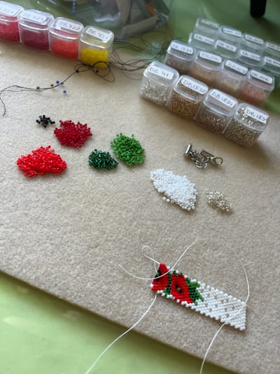 This image shows a close-up view of materials for beadwork, likely in Seredivka, Ukraine. The main subjects are various small beads in different colors, including red, green, black, white, yellow, and metallic tones, stored in small clear plastic containers with labels. Some containers have labels that appear to be product codes, such as "DB 1734," "DB 1832," "DB 1831," "DB 421," "DB 422," and "DB 423."
In the center of the image, there's a partially completed beadwork piece, which appears to be a bracelet or a bookmark, featuring a pattern of red poppies with green leaves on a white background. Nearby, there are small piles of loose beads in various colors, including red, green, and white, as well as a silver-colored clasp with a chain. A piece of white thread is visible, indicating that the beadwork is in progress. The beads and materials are laid out on a textured, light-colored surface, possibly felt or a similar fabric, with a hint of a green surface in the background. The lighting suggests an indoor setting, possibly a craft room or a workspace, with no clear indication of the time of day or weather. The overall impression is one of a hobby or craft activity, with the focus on the detailed work of bead weaving.
