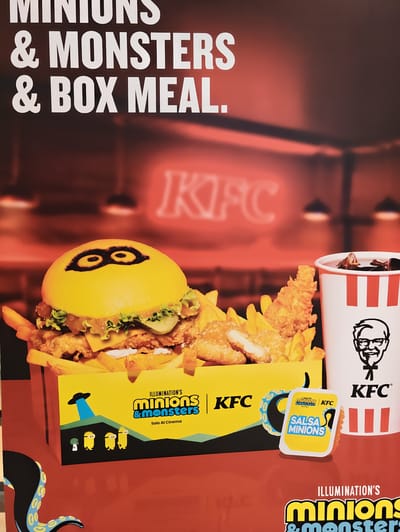 This is a close-up, eye-level promotional image of a KFC meal box and drink, likely for a movie tie-in. The foreground features a KFC bucket filled with fried chicken pieces and french fries. To the left of the bucket is a burger with a yellow bun decorated to resemble a Minion's goggles. A small dipping sauce container, also with Minion branding, is placed in front of the KFC bucket. The background shows a blurred, red-lit interior with a neon KFC sign. The overall aesthetic is vibrant and promotional, geared towards families and fans of the Minions franchise. There are no people depicted in the image, and no specific location in Nola, Italy is mentioned or visually identifiable.