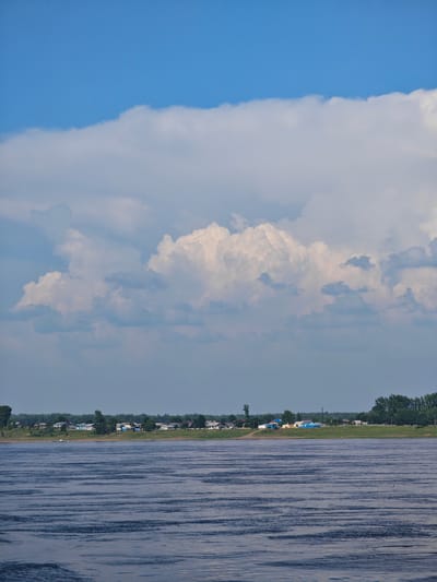 The image depicts a wide river in the foreground, with gentle ripples on its surface. The water appears deep blue. In the background, a village is situated on a grassy embankment. The village consists of several houses with colorful roofs, predominantly blue and white, interspersed with green trees. Beyond the village, a dense line of trees forms a dark green backdrop. Above the landscape, a vast sky dominates the upper portion of the image. This sky is characterized by a clear blue expanse in the upper left, transitioning into dramatic, voluminous white and gray cumulus clouds that stretch across the horizon. The overall scene suggests a tranquil rural setting in Verkhnepashino, Russia, likely on a summer day given the lush greenery and cloudy sky. There are no people or animals visible in the frame.
