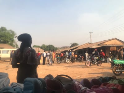 The image depicts an active outdoor market or street scene in Kwal, Nigeria, under a clear, bright sky. In the immediate foreground, a display of assorted colorful plastic footwear is visible. A person, viewed from behind and indistinct, stands in the left midground, observing the activity on the unpaved street. This thoroughfare is populated by numerous individuals and motorcycles. A white van is partially visible on the left. Along the right side of the street, a row of single-story buildings with corrugated metal roofs suggests market stalls or shops. Several men are present, including one in a red cap riding a red motorcycle towards the right. Another individual on the far right carries a yellow container on their head. A prominent utility pole with multiple overhead wires stands on the right. The ground is entirely unpaved dirt. Distant trees and a clear horizon form the background.