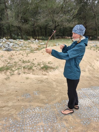 The scene depicts a fair-skinned adult individual standing outdoors in Thuận An Ward, Vietnam. The individual, seen in profile facing left, is wearing a blue and white patterned beanie, a light beige knit scarf, a teal hooded rain jacket, and black pants. Black thong sandals are on their feet, which are otherwise exposed. They are holding a small, unvarnished wooden bow with their left hand, aiming forward. A thin, light-colored string is strung on the bow, secured with a small pink or red tie near the upper limb. Their right hand is positioned near the bowstring, suggestive of drawing or preparing to draw.

The foreground consists of light brown sand partially covering an irregular grey paved or cobblestone surface. Scattered patches of sparse green vegetation are present in the sandy areas. The mid-ground features several piles of light-colored, angular rocks on the sand. The background is dominated by a dense line of tall, slender trees with dark green foliage, consistent with pine or casuarina trees, under an overcast sky.