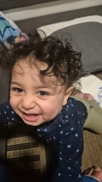 One infant is depicted in a close-up, indoor setting. The infant has dark, curly hair and dark eyes, and is smiling broadly, exposing gums. They are wearing a dark blue long-sleeved shirt with light-colored star patterns and light green trousers, partially visible. The infant is positioned on what appears to be a dark-colored rug with light horizontal stripes, looking directly at the camera. The background consists of soft furnishings, including a dark grey upholstered surface, a multi-colored patterned fabric on the left, and a white patterned fabric with a subtle blue design on the right. The specific location within the city of Melle, Belgium, is not discernible from the provided image.