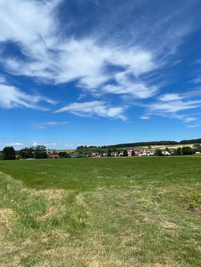 A landscape photograph shows a rural village nestled on a gentle slope under a bright blue sky dotted with white, wispy clouds. In the foreground, a vast, green field dominates the lower half of the image, with slightly drier patches and tire tracks suggesting agricultural use. The middle ground reveals a cluster of houses with red-tiled roofs and white walls, forming a village that extends along the curve of the land. Trees and vegetation surround the village, and a dark green line of forest borders the horizon. To the far left, a large, dense tree stands prominently. The overall scene depicts a peaceful, sunlit day in a German countryside setting, likely Christerode, Germany, given the context. There are no people or animals visible in the frame.