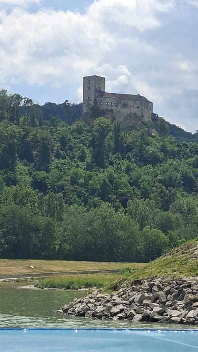 The image shows a medieval castle perched atop a hill covered in lush green trees. The castle has a prominent square tower and a long, low building attached to it. The sky above is partly cloudy, with patches of blue visible. In the foreground, a body of water, possibly a river, is visible, with a rocky shoreline on the right. The overall scene conveys a sense of historical grandeur and natural beauty. This is a photograph of the Forchtenstein Castle in Sankt Andrä-Wördern, Austria.
