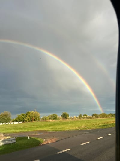 The image shows a double rainbow in Chascomús, Argentina. The view is from inside a vehicle. To the right, a paved road with white lane markings runs alongside a grassy field. The field has trees, bushes, and utility poles. In the sky, a prominent rainbow arc is visible, with a fainter secondary rainbow above it. The sky is overcast with gray clouds.