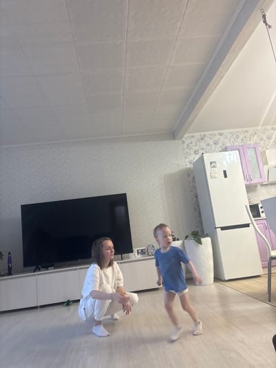 The image shows the interior of a room. A person wearing white clothing is squatting on the floor. A young child in a blue shirt and white socks is running. A black television is against a wall with wallpaper. A white refrigerator is near a white planter. There is light-colored wooden flooring. The specific location in Nestiukovo, Russia cannot be determined from the image.