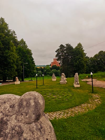The image depicts a park-like setting with several stone sculptures placed on a grassy lawn. In the background, a large, red-brick church with white domes and gold accents is visible. The sky is overcast, and the surrounding area is filled with lush green trees. A paved path winds through the grass, and there are several lampposts scattered throughout the scene. The foreground features a close-up of a blurred stone sculpture, suggesting it is a significant element of the park's display. The overall atmosphere is serene and somewhat somber due to the cloudy weather. This scene is located in Khotkovo, Russia, likely at the Museum-Estate Abramtsevo, a renowned art museum and cultural heritage site.