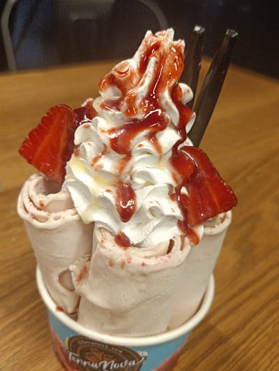 This is a close-up, eye-level, indoor shot of a dessert served in a paper cup. The dessert appears to be a rolled ice cream, with a strawberry flavor, topped with whipped cream and strawberry sauce. Two strawberry slices are visible on the side of the dessert, and two dark sticks, possibly for eating, are inserted into the whipped cream. The dessert is placed on a wooden surface. The background is dark and out of focus. The paper cup has a blue and red design with text that appears to read "Terranova Kara" and "Terra Nova". Based on the context of the cup, this dessert is likely from a "Terra Nova" establishment in Santa Teresa del Tuy, Venezuela.