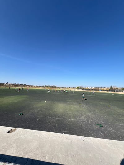 The image displays an outdoor artificial turf football field located in La Venta, Mexico, under a clear, bright blue sky. Approximately 20-30 individuals, primarily children or young adults, are scattered across the dark green and black turf. They are dressed in athletic wear, including shirts of various colors such as red, green, blue, and white, and appear to be engaged in a training or practice session. Several yellow training cones are distributed across the field, and a green fabric band lies on the turf in the mid-right. White line markings are faintly visible on the field. The background consists of a dry, sparsely vegetated landscape with distant low-rise structures and fences under a clear horizon. The foreground features a light grey concrete structure, possibly a barrier or spectator area, with a dark, irregularly shaped hole present on its top surface on the left side. The scene is illuminated by direct sunlight.