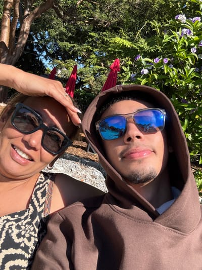 A woman and a man take a selfie outdoors. The woman wears black sunglasses, has red devil horns, and her hand is on her forehead. The man wears a brown hoodie and blue mirrored sunglasses. Vegetation, including a tree, is in the background. The sky is visible through the branches.