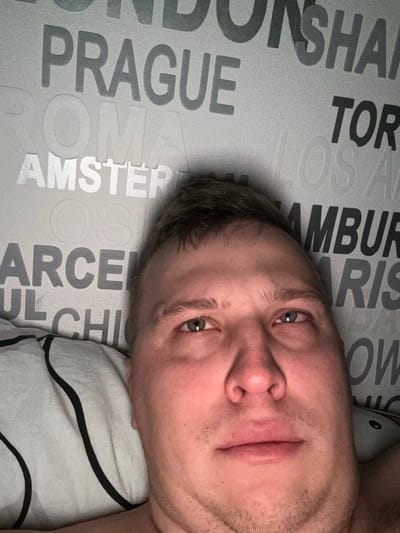 A man's face is captured close-up. His skin tone is fair, with visible stubble. He has short, light-brown hair and green eyes. He is lying on a pillow with black stripes on a white background. The background features a gray surface covered in the names of cities like London, Prague, Amsterdam, and Barcelona in different shades. The image is slightly rotated.