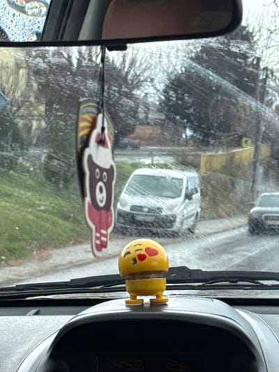 The image depicts a street scene in Kumanovo, North Macedonia, viewed from inside a vehicle through a wet, streaked windshield. In the immediate foreground, a yellow bobblehead ornament, styled as a kissing emoji with heart eyes, is affixed to the dashboard. Below it, a digital display shows '14:58'. A flat, hanging ornament resembling a brown bear with a red scarf and white beanie is suspended from the rearview mirror.

Outside the vehicle, a wet, paved street is visible. A white panel van is parked or stationary on the right side of the street, its front partially obscured by the windshield's condition. A dark-colored vehicle is partially visible behind the white van. To the left of the street, a grassy verge runs parallel to the pavement. The background features bare or sparsely foliated deciduous trees and a low, yellowish-beige wall or building segment on the right. The sky is overcast.