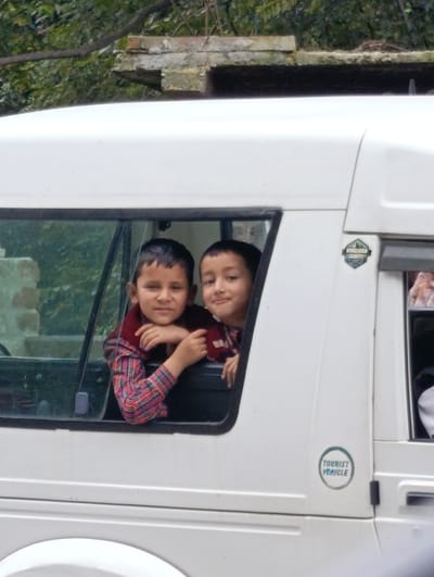 Two young boys are visible looking out of the window of a white van. The boy on the left has his arms resting on the window ledge, and the boy on the right is leaning forward slightly. Both boys appear to be smiling. The van has a "TOURIST VEHICLE" sticker on its exterior. The background suggests an outdoor setting with greenery and a structure above the van, possibly indicating a location in Gadherni, India, a region known for its natural beauty.