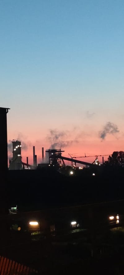 The image depicts an industrial landscape at dusk, likely in Salzgitter, Germany, given the context of industrial facilities. The sky transitions from a clear blue at the top to a soft pink and orange gradient near the horizon, indicating sunset or sunrise.
Prominently featured in the mid-ground are structures of a steelworks or similar industrial plant. Tall, silhouetted chimneys and towers rise against the colored sky. Smoke or steam is visibly emanating from some of these structures, creating a hazy effect. Various platforms, conveyor belts, and cranes are discernible, forming the complex architecture of the industrial site. Artificial lights are visible across the structures, some shining brightly, others appearing as scattered points of illumination.
In the foreground, silhouetted against the industrial scene, is a dark area that appears to be a collection of buildings or structures, possibly residential or other urban elements. Some rectangular lights, possibly from windows or streetlights, are visible within this foreground area, suggesting a human presence or activity despite the dim lighting. The very bottom of the image shows a dark, textured surface that might be a fence or railing. The overall impression is one of an industrial city at the transition between day and night, with human activity juxtaposed against a large-scale industrial environment.