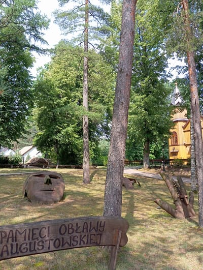 In Mikaszówka, Poland, an outdoor scene unfolds with a focus on artistic installations within a natural setting. A wooden sign in the foreground reads "PAMIĘCI OBLAWY AUGUSTOWSKIEJ". To the left, a large, carved wooden head sculpture rests on the ground, its features weathered and indistinct. In the middle ground, to the right of the head, a large, stylized metal sculpture of a hand, adorned with beads and a cross, is partially embedded in the grass. The hand appears to be part of a larger conceptual artwork. Behind these sculptures, a pathway winds through a grassy area with scattered trees. In the background, a traditional wooden church with a steeple, painted yellow, stands prominently. Several white buildings, possibly houses, are visible further to the left, nestled amongst trees. The environment is characterized by a dense canopy of green trees, with sunlight dappling the ground, suggesting a warm, possibly summer day.