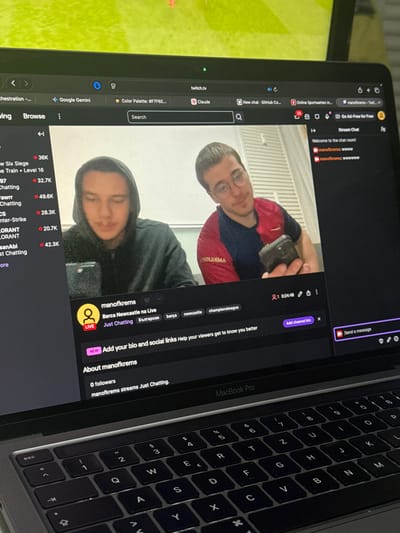The image displays a MacBook Pro laptop screen open to twitch.tv, featuring a live stream from a channel named "manofkrems." This channel name suggests a connection to Krems an der Donau, Austria. The stream is titled "Barca Newcastle na Live" and is categorized as "Just Chatting," with tags including "български," "barça," "newcastle," and "championsleague."
The stream video shows two young adult males. The individual on the left wears a dark hooded sweatshirt and is looking downwards. The individual on the right, wearing a red and dark blue two-toned shirt and glasses, smiles faintly towards the camera while holding a small, dark, rectangular electronic device in his right hand. The background behind the streamers is a plain, light-colored wall with vertical indentations.
On the right side of the Twitch interface, the stream chat is visible, displaying "Welcome to the chat room!" and subsequent messages from "manofkrems." The channel currently shows "0 followers" and the stream duration is approximately 24 minutes and 48 seconds. Above the Twitch page, several browser tabs are open, including "Google Gemini," "Color Palette: #F7F6E...," and "Claude." Partially visible above the laptop screen is a brightly lit, out-of-focus green surface, likely another monitor.