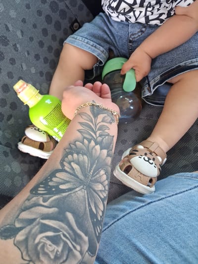 In Kukorevo, Bulgaria, a close-up, eye-level, outdoor, natural light shot shows a person's tattooed arm and a child's legs. The person's arm, which is to the left, features a large, grayscale tattoo of a rose and a butterfly. A thin, gold chain bracelet adorns the person's wrist. To the right, a child is wearing denim shorts and tan sandals with a tiger design. The child is holding a clear bottle with a green lid and the person is holding a bright green bottle. The background is a dark, patterned fabric, possibly from a car seat.