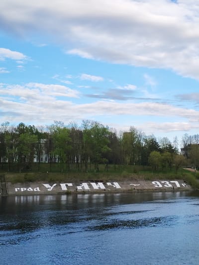 The image shows a view of the Kotorosl River in Uglich, Russia. On the riverbank, a concrete embankment is painted with the inscription "Град Углич - 937 г." which translates to "City of Uglich - 937 AD." Trees line the embankment behind the concrete structure. The sky above is blue with scattered white clouds. The water appears slightly choppy. Two people are visible along the inscription on the bank.