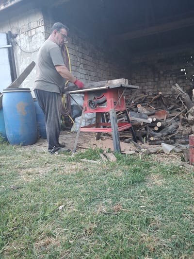 This media file features a man standing in an outdoor, rustic setting. He is wearing glasses, a grey t-shirt, dark pants, and red gloves. The man is positioned next to a red table saw with a circular blade. He appears to be engaged in a task involving the saw and some wooden materials. The background is a jumble of cut wood and what looks like a partially constructed brick structure. To the left, there's a dark, shadowy area, possibly an open shed or building. To the right and in the foreground, there's a patch of grass, some of which is dry and yellowish, suggesting it might be a rural or backyard area. Two large blue barrels are visible in the lower right portion of the image, one of which is partially obscured by the man's legs. The overall impression is one of a DIY project or outdoor work. The lighting suggests it is daytime, and the weather appears to be fair. The scene is set in Kochmar, Bulgaria. Visible text on the table saw includes "Einhell".