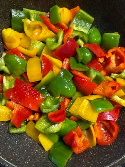 A close-up, overhead shot shows chopped bell peppers in a dark pan. The peppers are a mix of red, green, yellow, and orange, with some pieces showing slight charring. The pan appears to be non-stick with a speckled dark gray surface. The lighting is artificial and casts a slight sheen on the peppers. The image does not contain any people or specific environmental context to determine a location in Southbank, Australia.