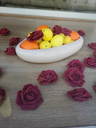 The image depicts a still life arrangement indoors, set on a surface resembling light wood flooring. A shallow, elongated, speckled white bowl is centrally positioned, containing an assortment of fruits – oranges and lemons. Dark red artificial roses are scattered both within and around the bowl, creating a decorative effect. The arrangement appears to be in front of a window, which is partially visible in the background. There is no indication of the location being specifically in the city of Pardubice, Czechia.